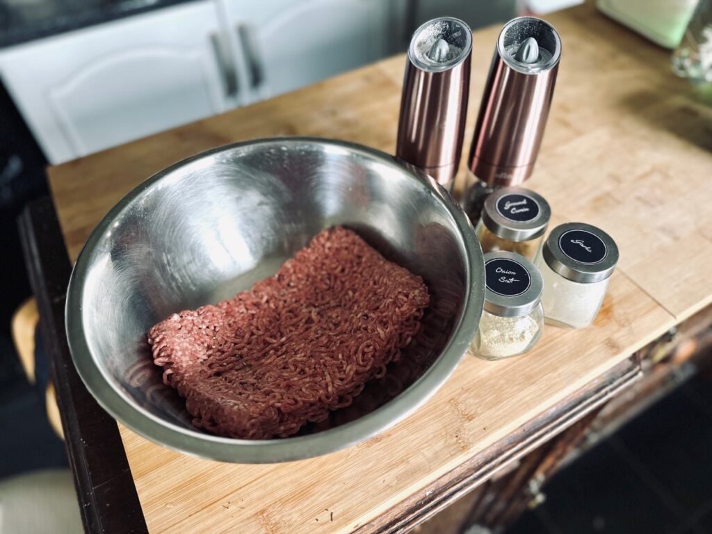 A steel bowl containing mince meat, next ro spice jars and salt and pepper grinder on a wooden chopping board
