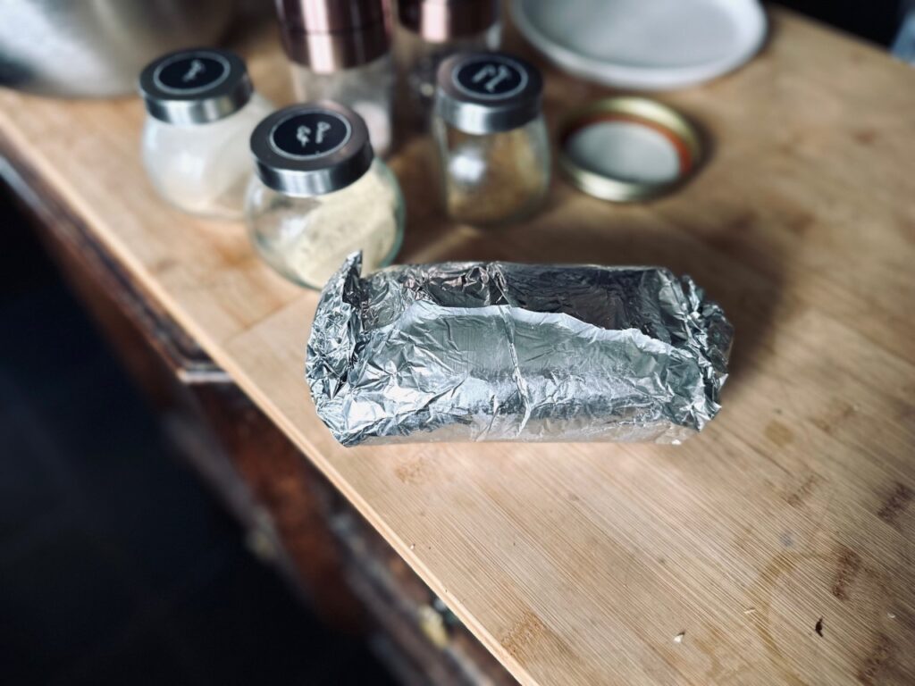 A foil package on a wooden chopping board with jars of herbs and spices behid