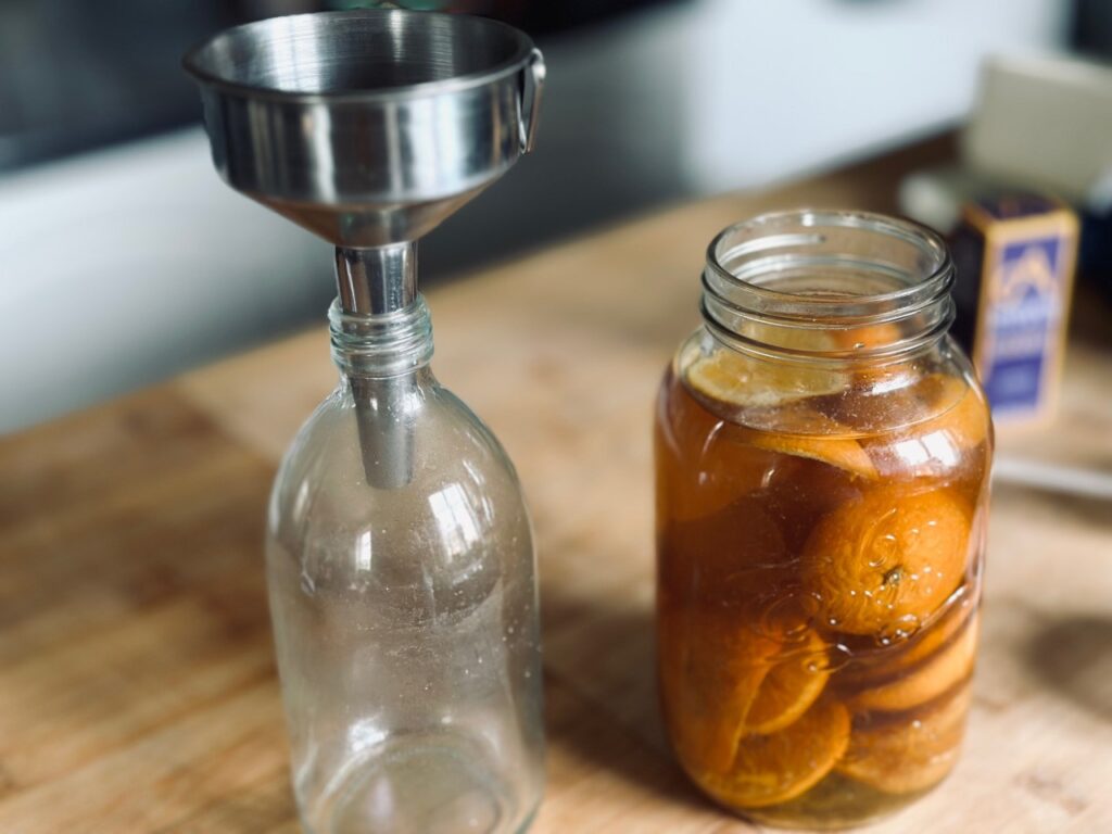 A glass bottle with a funnel next to a jar of oranges in vinegar on a wooden worktop