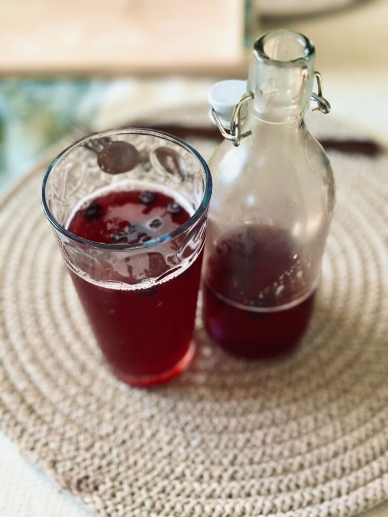 A glass of purple liquid next to an open bottle of Kombucha on a spiral placemat
