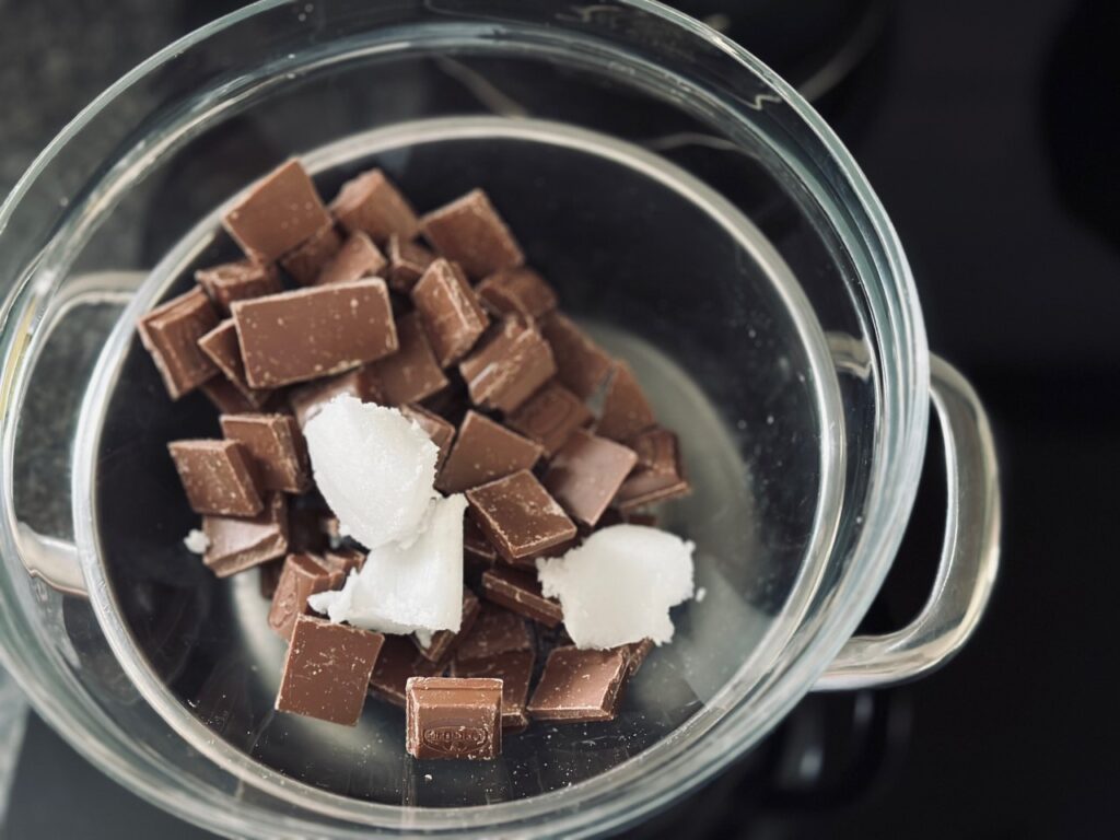 A glass bowl containing chocolate squares and dollops of solid coconut oil on top of a pan