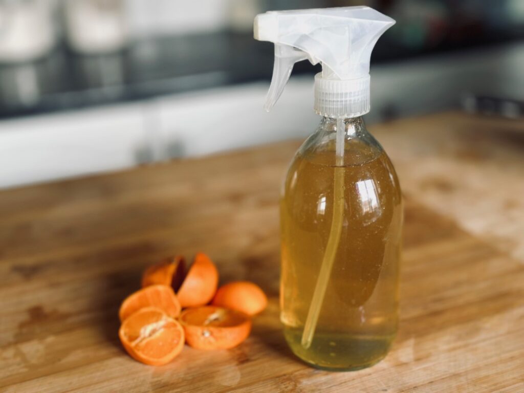 A glass bottle of orange natural cleaner on a wooden chopping board next to some sliced tangerine