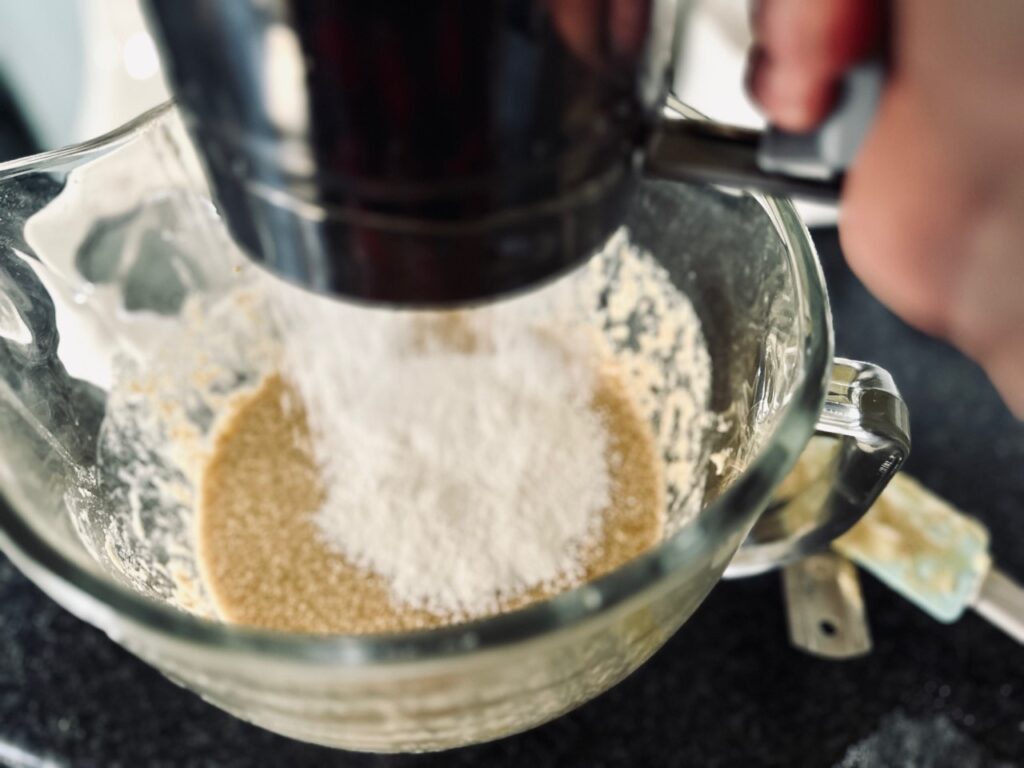 A glass bowl with batter and flour being sifted into it