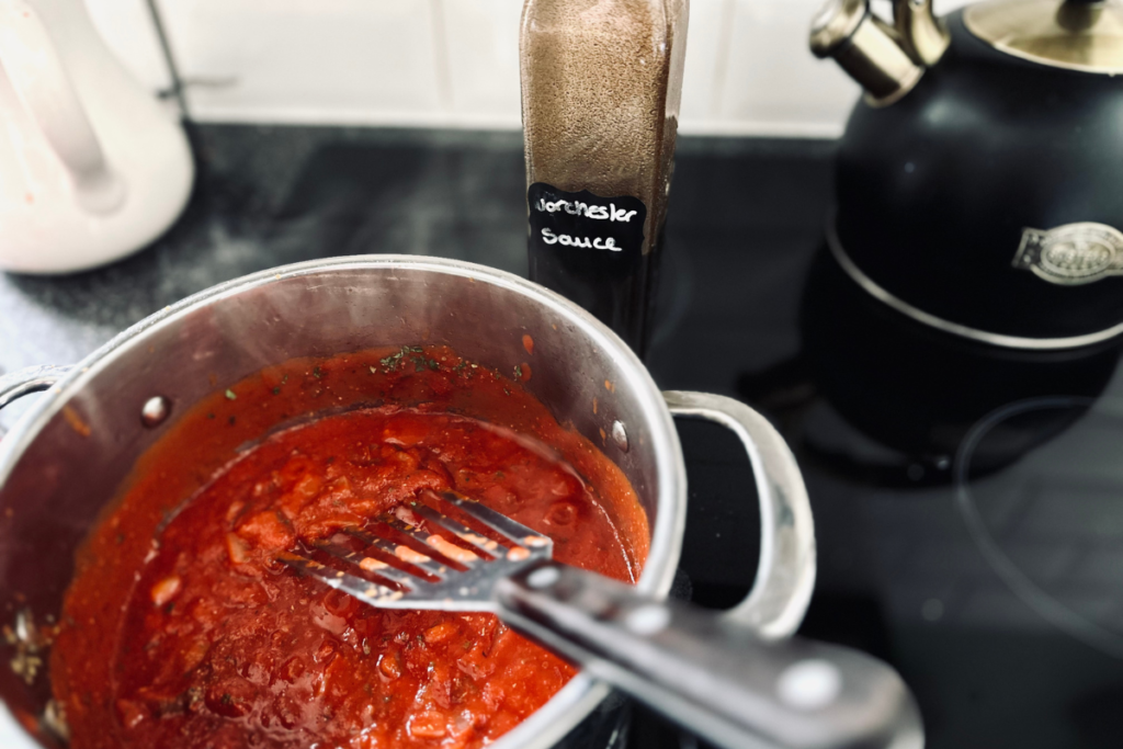 A saucepan with simple pasta sauce being stirred and a bottle of decanted Worcestershire sauce next to it