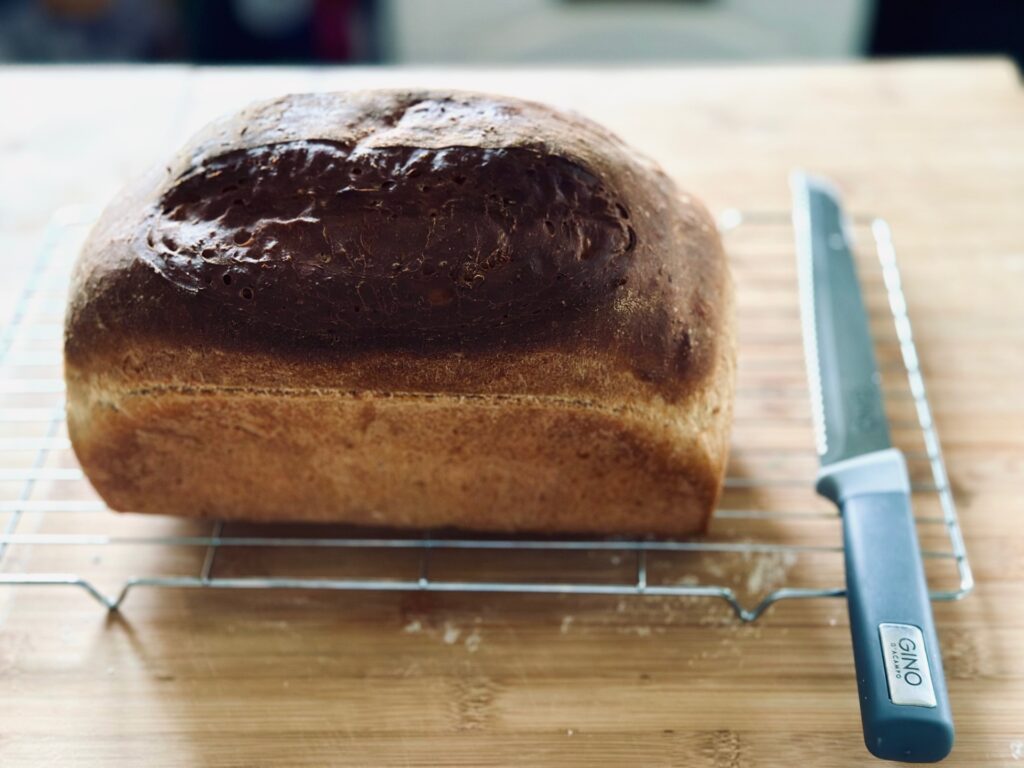 A cooked loaf of bread on a cooling rack with a bread knife beside it