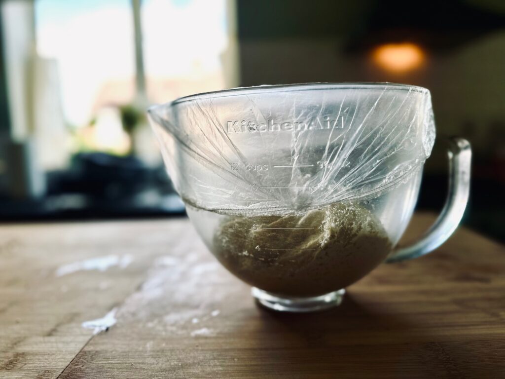 A side view of a mixing bowl covered in plastic, containing bread dough