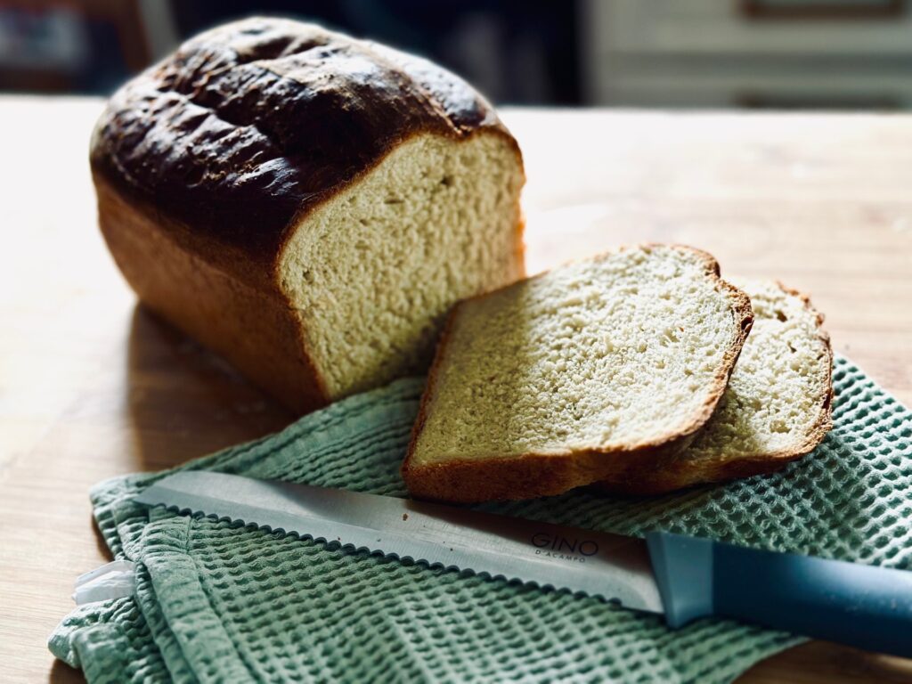 A loaf of bread with 2 slices cut from is, on a towel with a bread knife laid in front of it