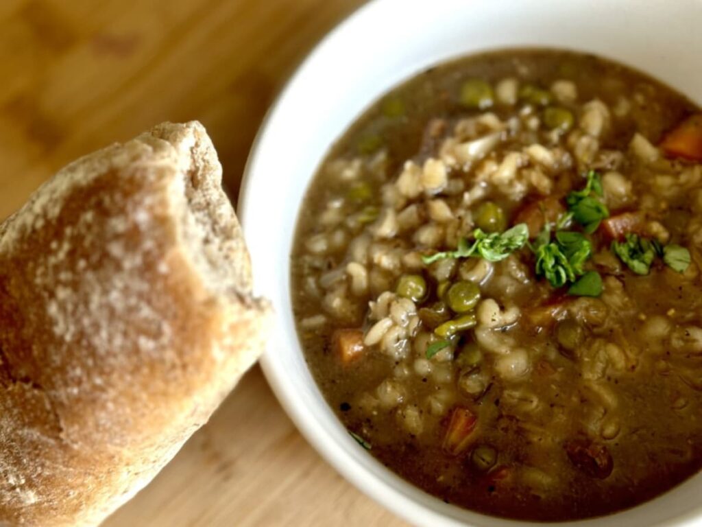 A bowl of slow cooker beef and barley stew with a bread roll beside it