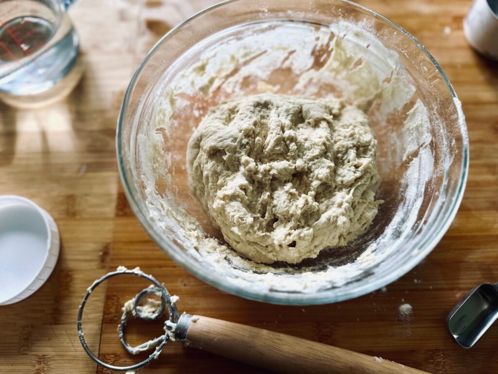 A bowl of shaggy bread dough and a dough whisk on a wooden board
