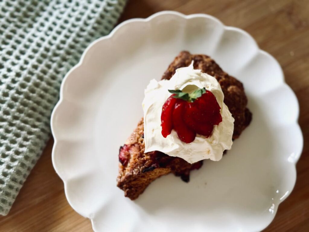 A sourdough discard scone topped with cream and a sliced strawberry sitting on a white plate
