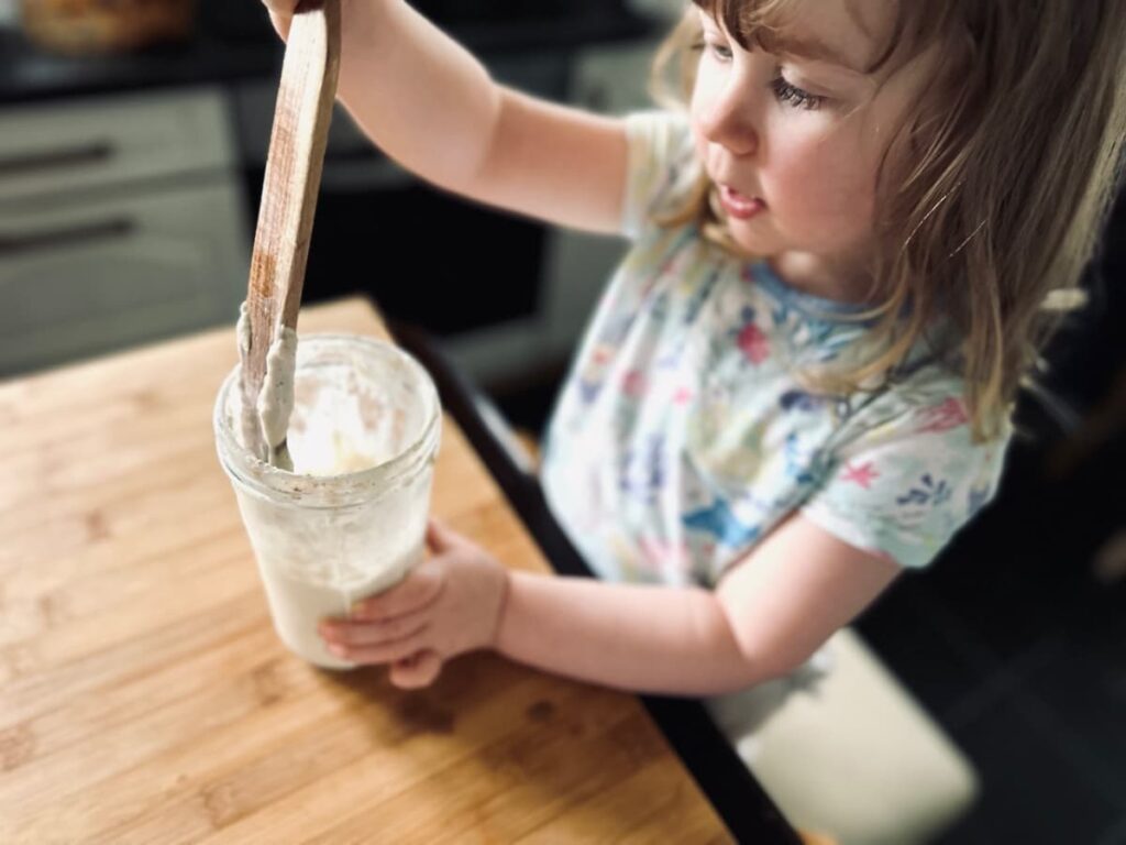 a little girl stood up at a counter stirring a jar of sourdough starter