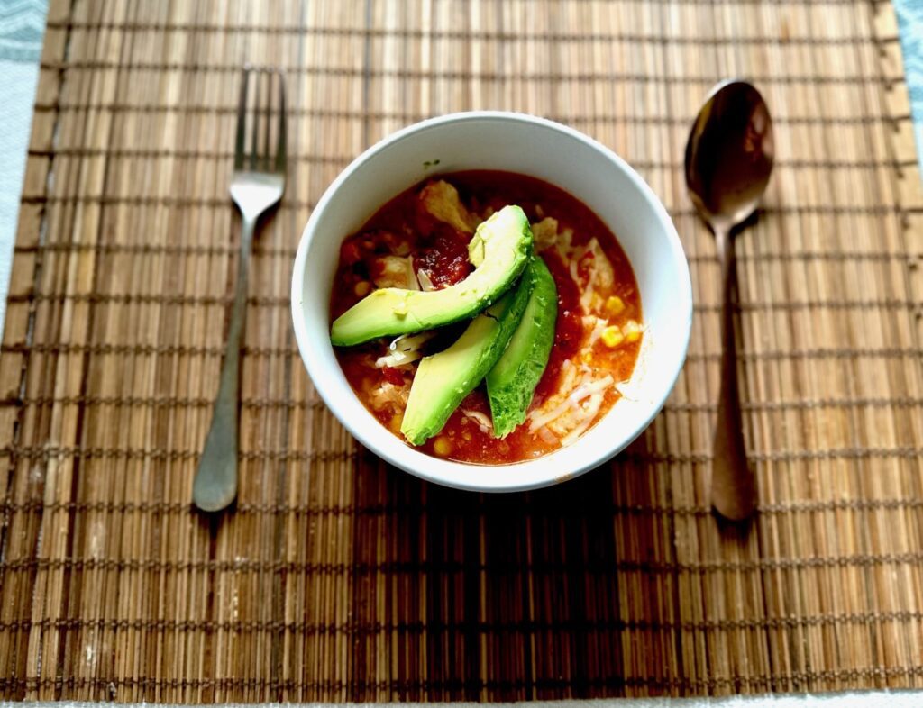 A bowl of Taco Stew topped with avocado, on a bamboo placemat with a spoon and fork beside it