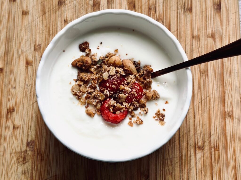 A white bowl of yoghurt containing granola and raspberries on a wooden chopping board