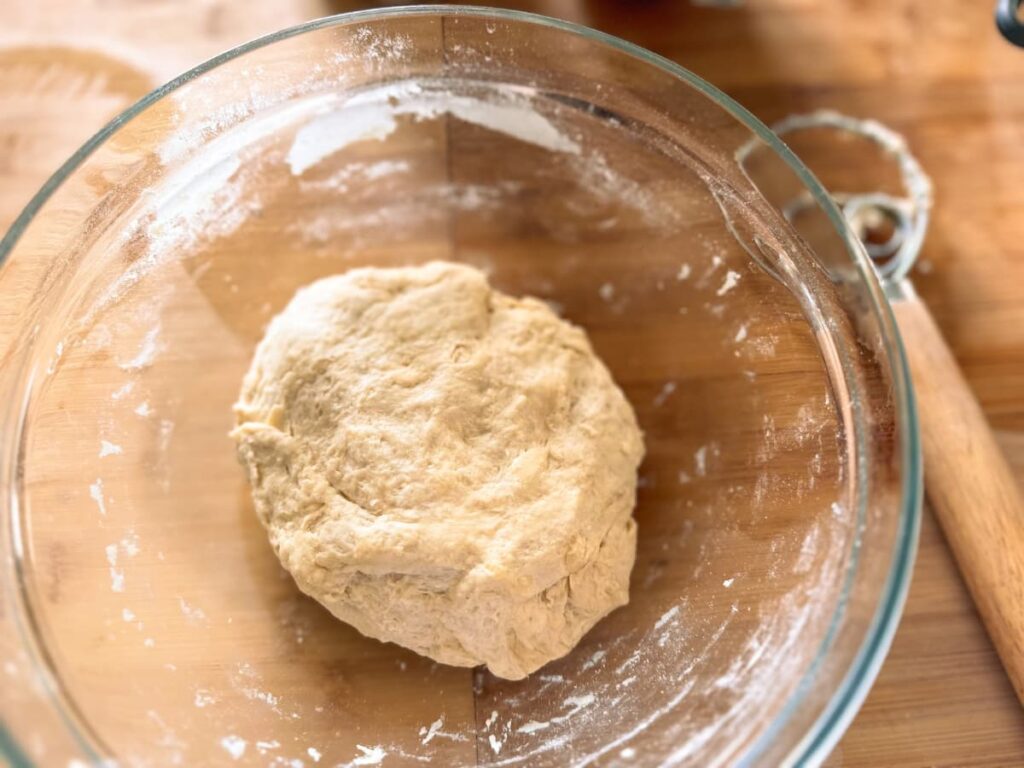 A glass bowl containing sourdough tortilla dough, with a dough whisk beside it