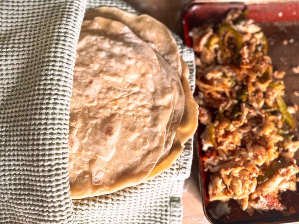 A pile of sourdough tortillas wrapped in a towel beside a tray of fish tacos