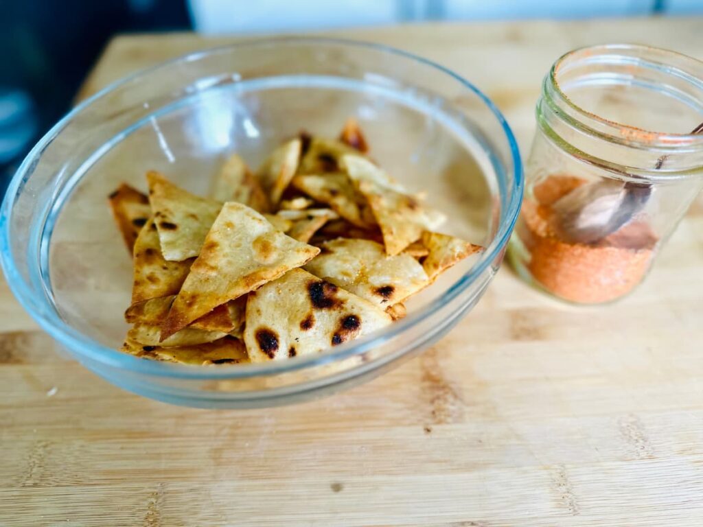 A glas bowl full of homemade tortilla chips next to a jar of taco seasoning with a spoon inside