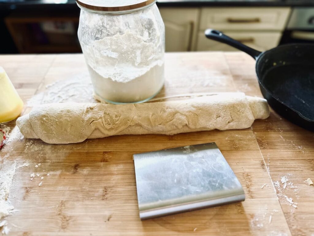 A rolled up log of lemon roll dough with a bench scraper and jar of flour
