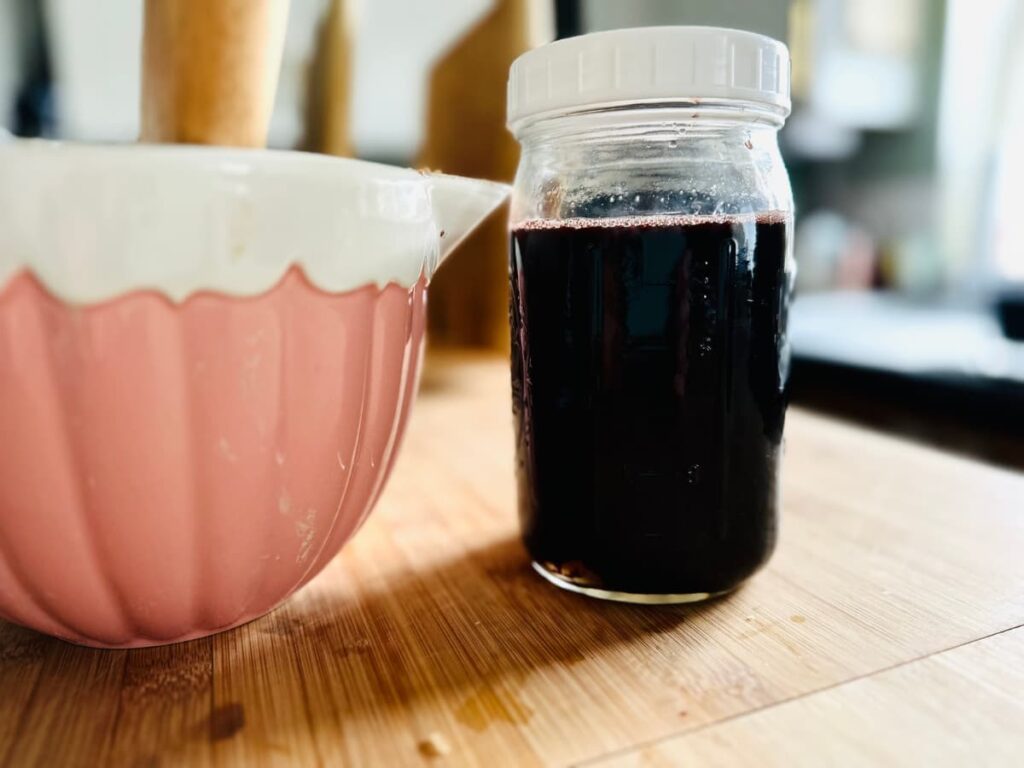 A jar of dark elderberry syrup next to a pink measuring jug