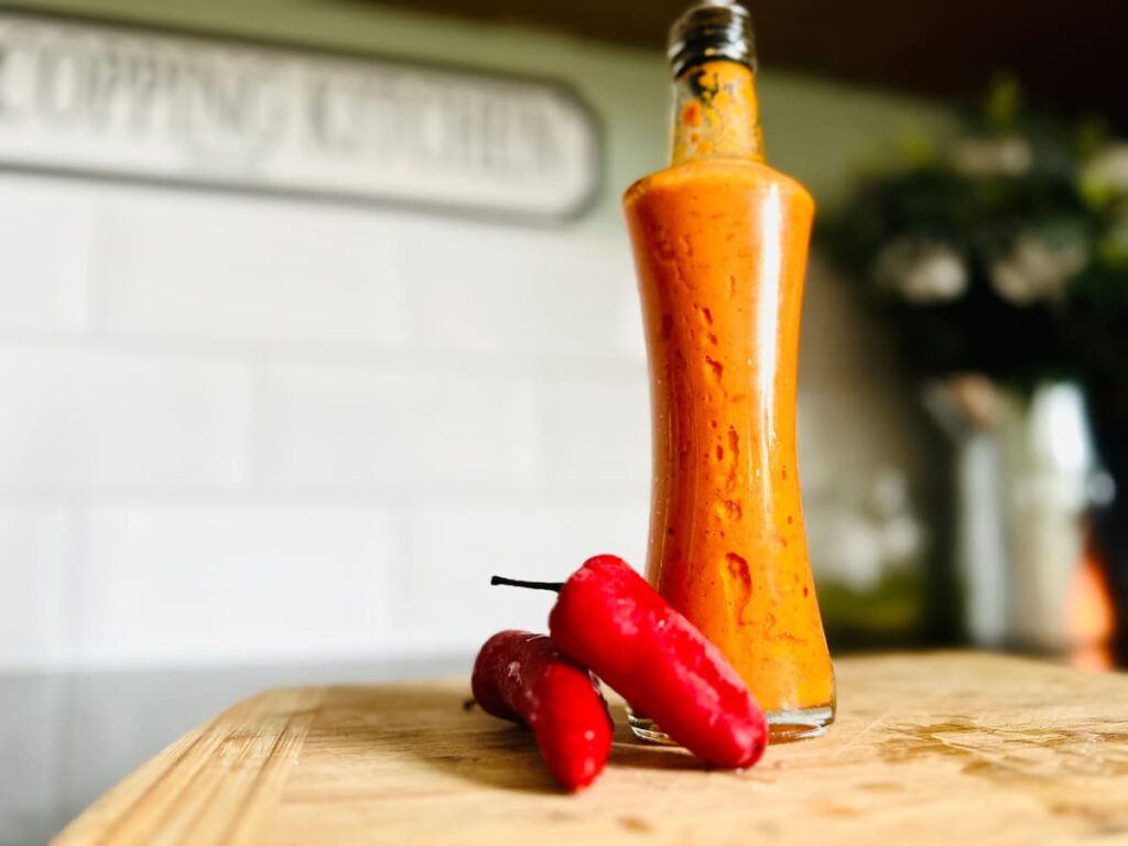 A glass bottle of orange coloured homemade Fermented Hot Sauce on a chopping board next to 2 red chillies
