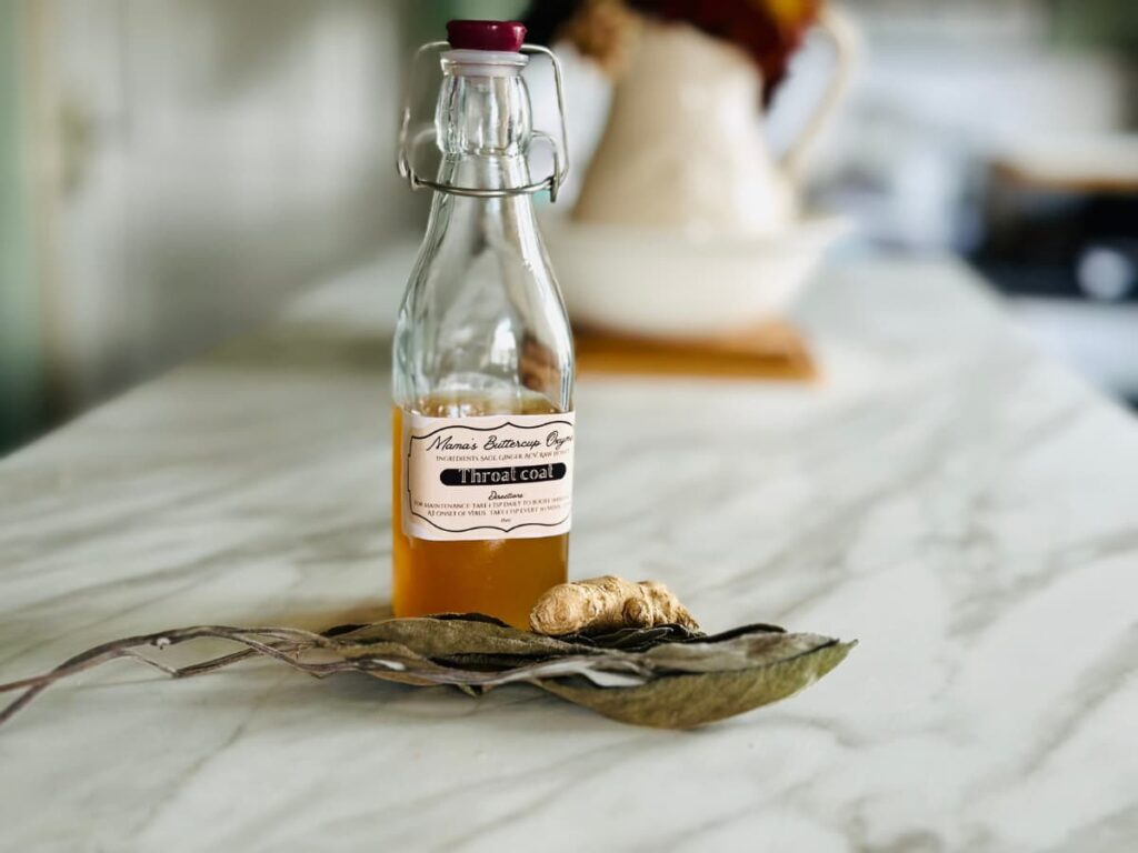 A bottle of amber liquid labelled Oxymel, beside dried sage leaves and ginger