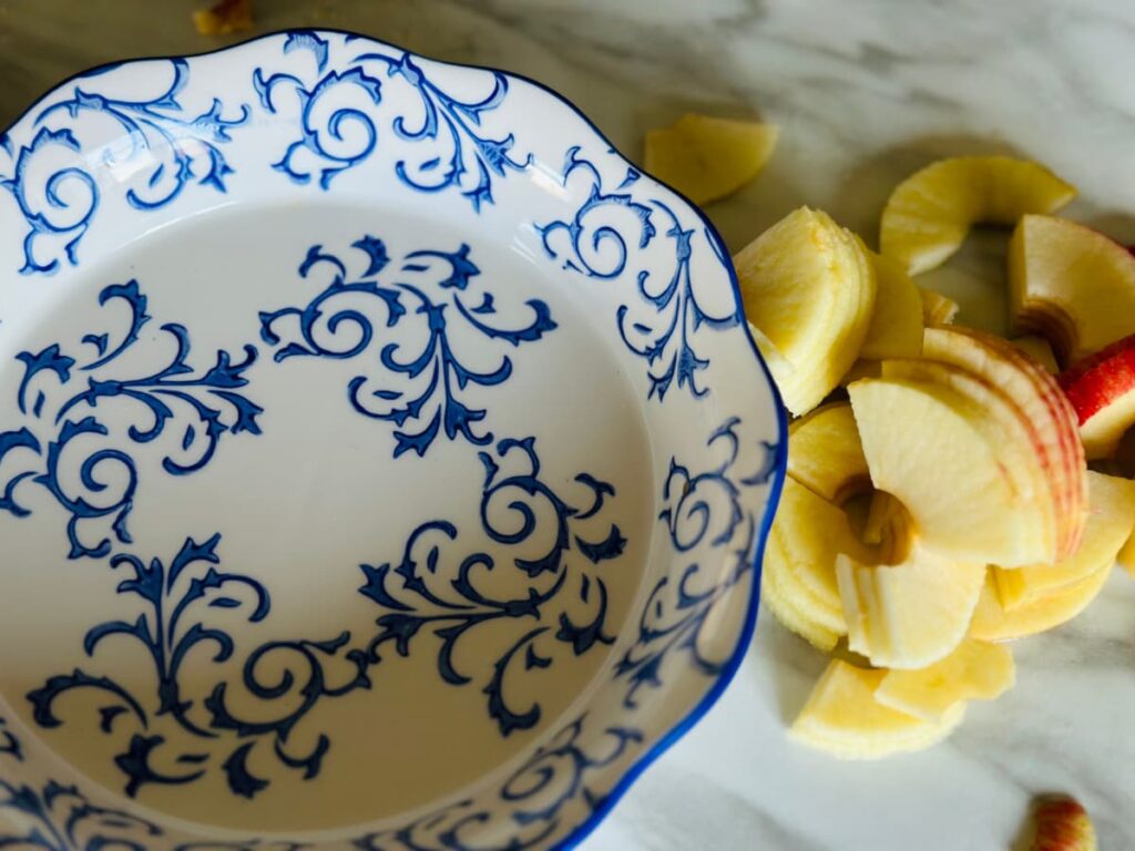 A decorative white and blue pie dish with chopped sliced apples next to it