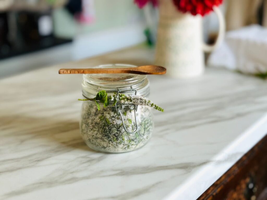 A jar of homemade herbal bath salts decorated with a mint flower and a spoon on top