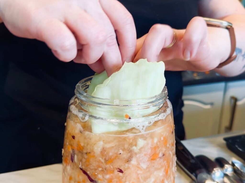 Someone placing a cabbage leaf into a jar of homemade Cortido to weigh it down
