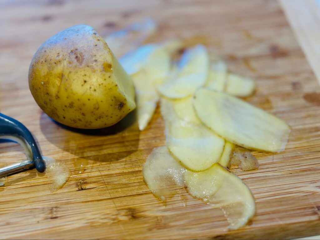 Thin slices of a potato next to a vegetable peeler and a half sliced potato